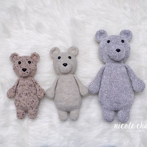 May include: Three crocheted teddy bears in different colors and textures. The bear on the left is brown with purple speckles, the middle bear is beige, and the bear on the right is gray.