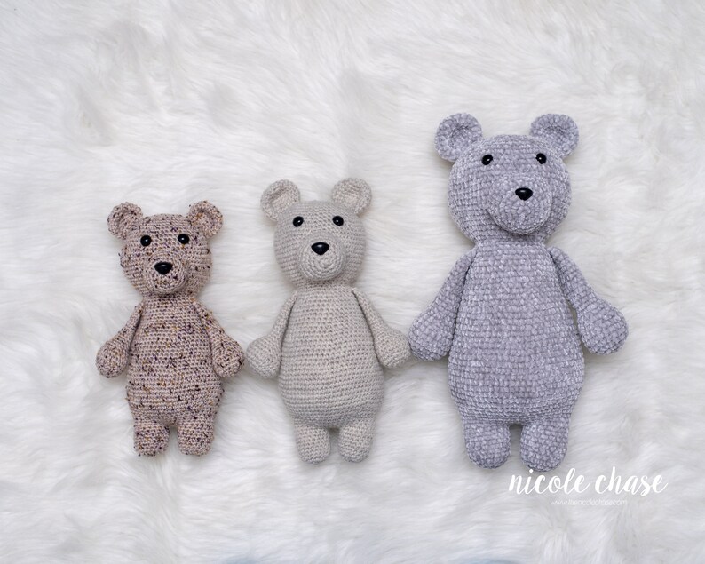 May include: Three crocheted teddy bears in different colors and textures. The bear on the left is brown with purple speckles, the middle bear is beige, and the bear on the right is gray.