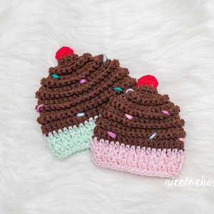 Peut inclure: Deux bonnets en crochet en forme de cupcake avec du gla&ccedil;age brun, des p&eacute;pites color&eacute;es et une cerise rouge sur le dessus. Un bonnet a un fond vert clair et l'autre un fond rose.