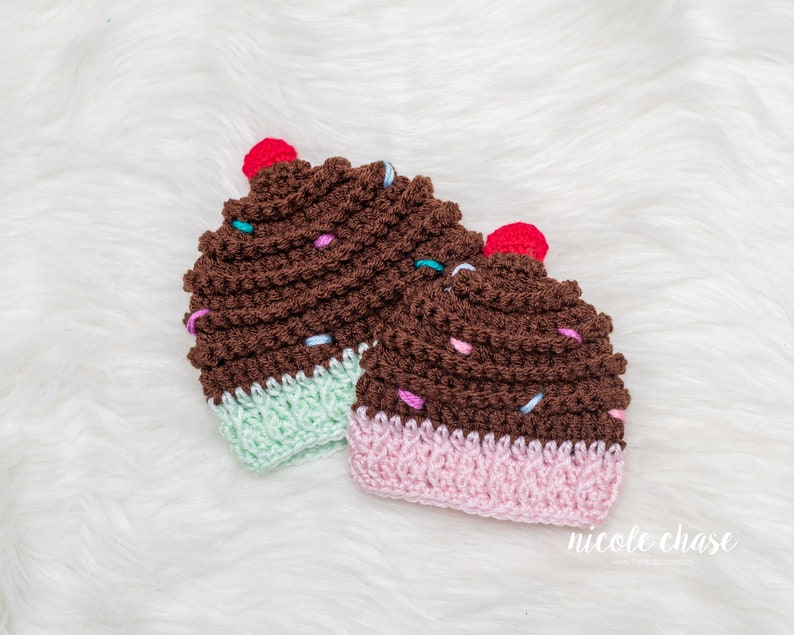 Peut inclure: Deux bonnets en crochet en forme de cupcake avec du gla&ccedil;age brun, des p&eacute;pites color&eacute;es et une cerise rouge sur le dessus. Un bonnet a un fond vert clair et l'autre un fond rose.