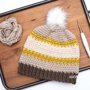 Pode incluir: Um chap&eacute;u de croch&ecirc; com um padr&atilde;o listrado marrom, bege e amarelo. O chap&eacute;u tem um pompom branco no topo e est&aacute; sentado em uma bandeja de madeira com um novelo de l&atilde; e um gancho de croch&ecirc;.