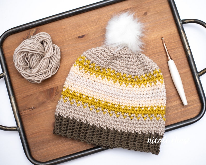 Pode incluir: Um chap&eacute;u de croch&ecirc; com um padr&atilde;o listrado marrom, bege e amarelo. O chap&eacute;u tem um pompom branco no topo e est&aacute; sentado em uma bandeja de madeira com um novelo de l&atilde; e um gancho de croch&ecirc;.