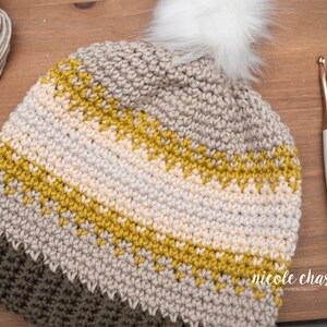 Pode incluir: Um chap&eacute;u de croch&ecirc; com um pompom branco e listras bege, amarelo mostarda e verde oliva. O chap&eacute;u est&aacute; sobre uma superf&iacute;cie de madeira com um gancho de croch&ecirc; ao lado.