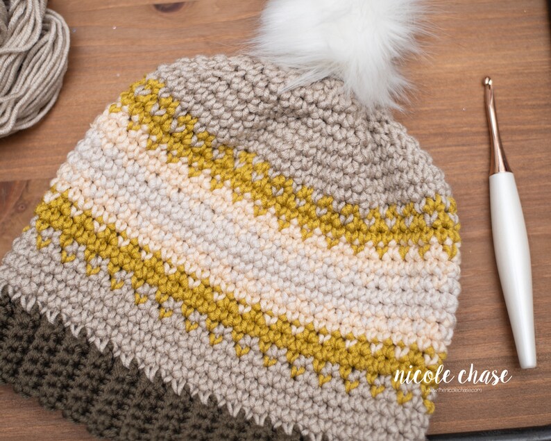 Pode incluir: Um chap&eacute;u de croch&ecirc; com um pompom branco e listras bege, amarelo mostarda e verde oliva. O chap&eacute;u est&aacute; sobre uma superf&iacute;cie de madeira com um gancho de croch&ecirc; ao lado.