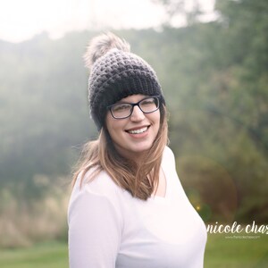 May include: A woman wearing a gray and black knit beanie with a faux fur pom pom, black framed glasses, and a white long-sleeved shirt. She is smiling and looking to the right. The background is blurred and out of focus.