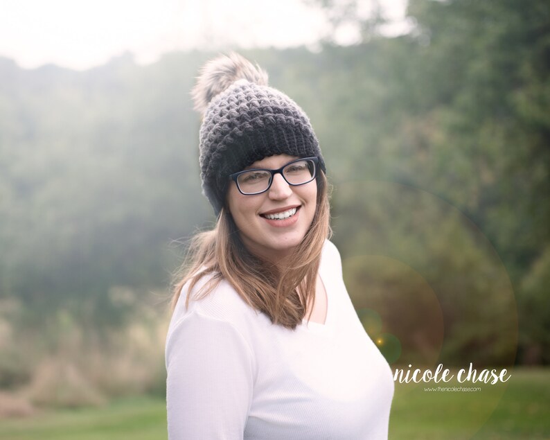 May include: A woman wearing a gray and black knit beanie with a faux fur pom pom, black framed glasses, and a white long-sleeved shirt. She is smiling and looking to the right. The background is blurred and out of focus.