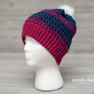 May include: A crocheted beanie hat with a pom pom on top. The hat is made with a combination of pink and blue yarn. The hat is on a white mannequin head.