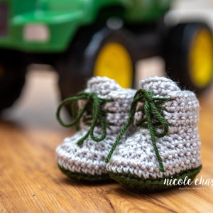 May include: A pair of gray crocheted baby booties with green laces and green trim. The booties are sitting on a wooden surface in front of a green toy tractor.