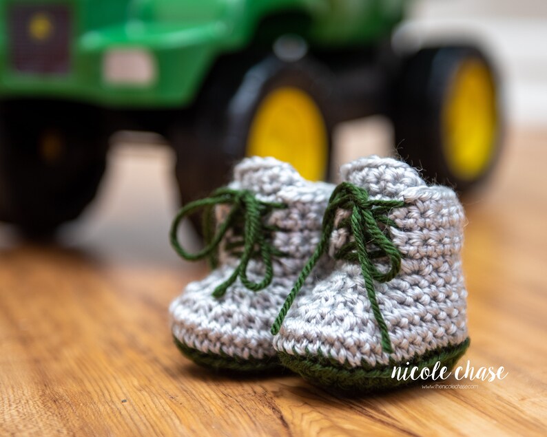 May include: A pair of gray crocheted baby booties with green laces and green trim. The booties are sitting on a wooden surface in front of a green toy tractor.