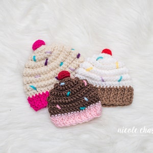 Peut inclure: Trois bonnets en crochet en forme de cupcake dans diff&eacute;rentes couleurs et designs. Les bonnets sont blancs, bruns et beiges avec un gla&ccedil;age rose, blanc et brun et des p&eacute;pites color&eacute;es.