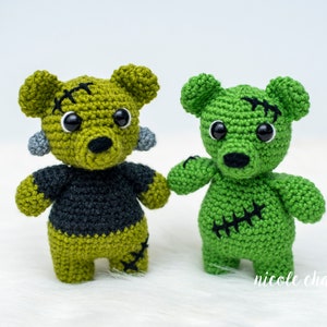 May include: Two green crocheted teddy bears with black eyes and stitched details. One bear has a black shirt and the other has green stitching on its chest.
