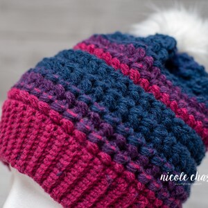 May include: A crocheted beanie hat with a white pom pom. The hat is striped with shades of pink, purple, and blue.