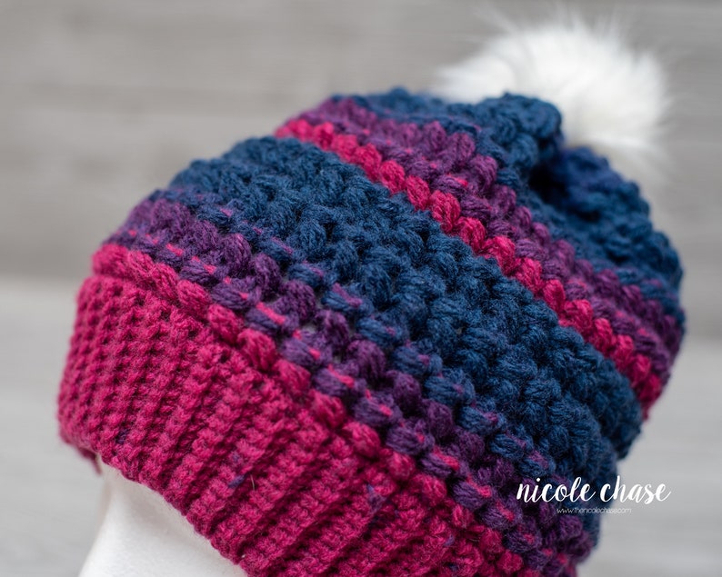 May include: A crocheted beanie hat with a white pom pom. The hat is striped with shades of pink, purple, and blue.
