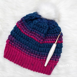 May include: A blue and pink striped crochet beanie with a white pom pom and a white crochet hook.