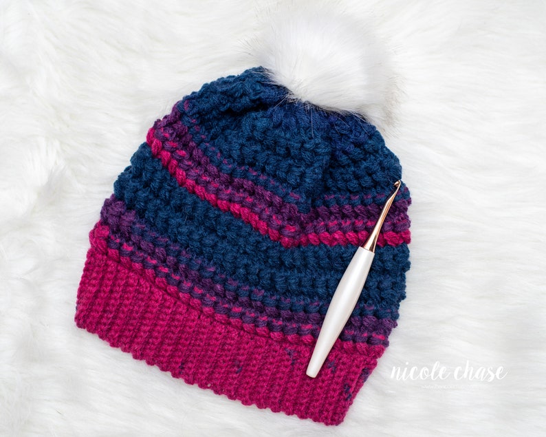 May include: A blue and pink striped crochet beanie with a white pom pom and a white crochet hook.