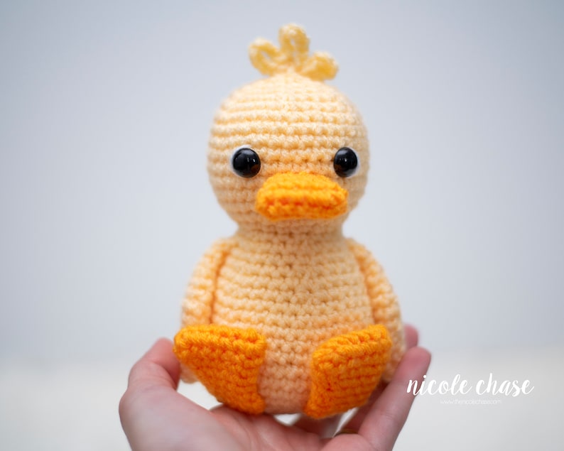 May include: A crocheted yellow duck with orange feet and beak. The duck has black eyes and is sitting in a hand.