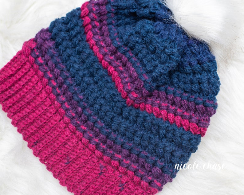 May include: A crocheted beanie hat with a striped pattern in shades of pink and blue. The hat is made with a textured stitch that gives it a puffy look.