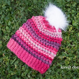 May include: A pink and purple crocheted beanie with a white pom pom. The beanie is lying on a bed of green grass.