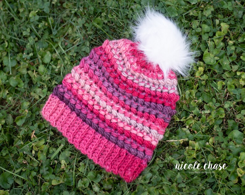 May include: A pink and purple crocheted beanie with a white pom pom. The beanie is lying on a bed of green grass.