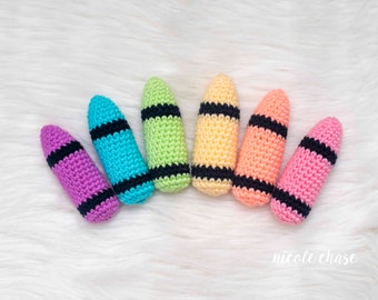 May include: A set of seven crocheted crayons in various colors, including purple, blue, green, yellow, orange, pink, and red. Each crayon has a black stripe around the middle.