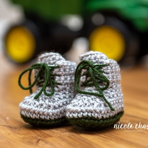 May include: A pair of gray crocheted baby booties with green laces and green trim. The booties are in focus, while a green tractor is blurred in the background.