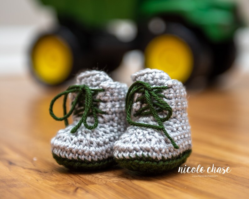 May include: A pair of gray crocheted baby booties with green laces and green trim. The booties are in focus, while a green tractor is blurred in the background.