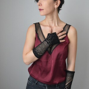 May include: A woman wearing a burgundy tank top with black lace detailing and black fishnet fingerless gloves. She is wearing dark wash jeans.