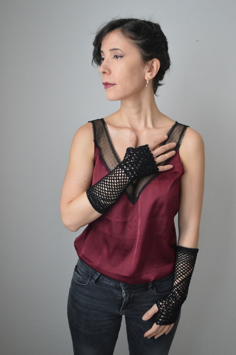 May include: A woman wearing a burgundy tank top with black lace detailing and black fishnet fingerless gloves. She is wearing dark wash jeans.