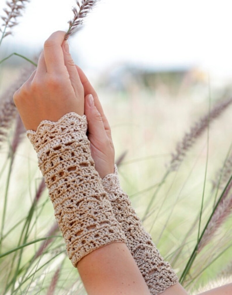 May include: A pair of crocheted wrist warmers in a light beige colour. The warmers have a delicate lace pattern and are shown against a blurred background of tall grass.