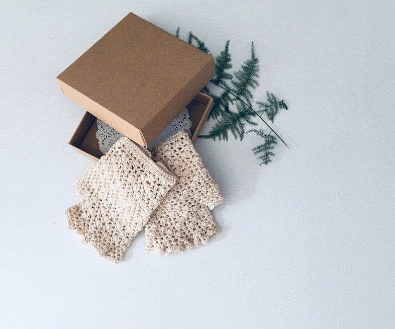 Peut inclure: Deux mitaines en crochet beige dans une bo&icirc;te en carton marron avec une dentelle blanche &agrave; l'int&eacute;rieur. Une foug&egrave;re verte est en arri&egrave;re-plan.