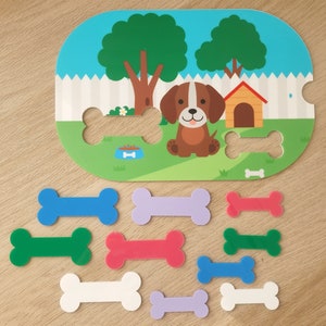 May include: A colourful wooden puzzle featuring a cartoon dog in a backyard setting. The puzzle includes nine pieces shaped like bones in various colours, including blue, green, purple, pink, and white.