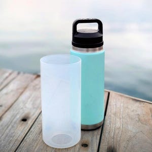 Puede incluir: Una botella de agua aislada de color turquesa con asa y tapa negras, junto a una funda protectora cilíndrica transparente. La botella tiene una base de acero inoxidable. Ambos artículos están sobre una superficie de madera, con un fondo de agua borroso.