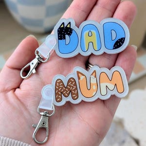 May include: Two keyrings, one saying "DAD" in blue with black details, the other saying "MUM" in orange and brown. Both keyrings have silver clasps. The keyrings are in a fun, cartoon style, perfect for gifting.