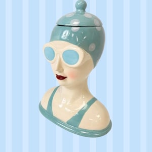 May include: A ceramic bust of a swimmer wearing a light blue swim cap with white polka dots and light blue goggles. The figure has a cream-coloured complexion, red lips, and a light blue swimsuit top. The head functions as a removable lid.