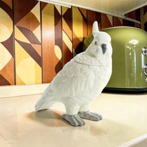 May include: A white ceramic cockatoo figurine with a textured feather pattern, a grey beak, and grey feet. The bird is depicted in a standing position with a crest on its head and a horizontal seam across the chest indicating a removable lid.