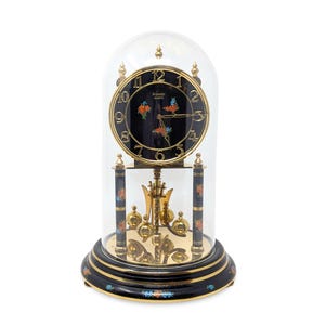 May include: A black Kundo Quartz clock with a glass dome. The clock face is black with gold numerals and hands, and floral accents. The base and columns are black with gold trim and floral designs. The clock has a pendulum and gold-colored weights.