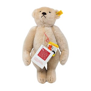 May include: A beige plush teddy bear with a dark brown stitched nose and mouth. A yellow Steiff ear tag is attached to the left ear. A white tag from The Margaret Woodbury Strong Museum and a yellow tag reading Für immer Dein are attached.