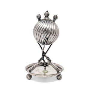 May include: A silver-colored, antique-style butter dish. The dish features a spherical, ribbed container with a decorative top, supported by crossed, ornate legs. It rests on a round base with three ball feet, showcasing a vintage aesthetic.