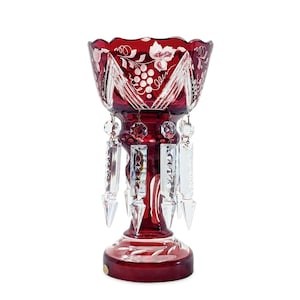 May include: A deep red cut-to-clear crystal vase featuring a scalloped rim, intricate etched grape and vine motifs, and a series of clear, faceted glass prisms suspended from the bowl by small metal rings, set on a circular pedestal base.