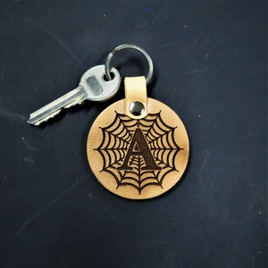 May include: A circular tan leather keychain featuring a dark brown laser-engraved spider web pattern with the capital letter A centered in the middle. The leather tag is attached to a silver metal split ring that holds a single silver house key.