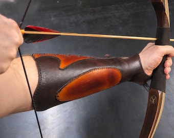 May include: A dark brown leather forearm guard with a lighter tan oval inlay, worn on the arm while holding a wooden recurve bow. The bow features a black grip and a small circular logo with a deer silhouette. An arrow with red fletching is nocked.