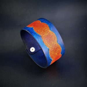 May include: A blue and orange leather cuff bracelet with a decorative floral pattern. The bracelet is made of thick leather and has a snap closure.