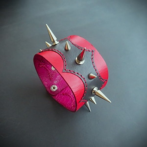 May include: A red and black leather cuff bracelet with silver spikes. The bracelet has a heart-shaped design with red stitching and a pink interior. The cuff is secured with two silver snaps.