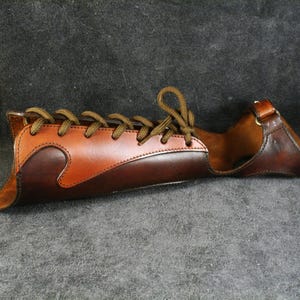 May include: A brown leather arm guard with a laced design and buckle closure. The leather is two-toned, with a lighter brown accenting the darker base. The guard is designed to protect the forearm and is suitable for archery.