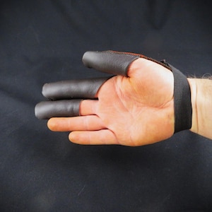 May include: A black leather finger tab for archery, with a wrist strap. The finger tab covers three fingers and is stitched with orange thread. The wrist strap is made of black elastic.