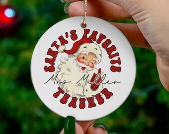 May include: White round Christmas ornament with a red and white Santa Claus illustration. The ornament says "Santa's Favorite Teacher" with the name "Mrs. Miller" written in cursive.