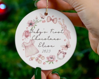 May include: A white ceramic Christmas ornament with a pink floral design. The ornament features the text "Baby's First Christmas Elise 2023".