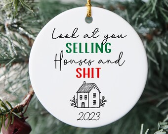 May include: A white ceramic Christmas ornament with a gold string, hanging on a Christmas tree. The ornament has the text "Look at you selling Houses and SHIT" with a house graphic and the year 2023.