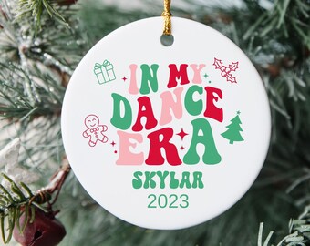 May include: White ceramic Christmas ornament with the text "In My Dance Era" in green, pink, and red letters. The ornament also includes a gingerbread man, a gift box, a holly sprig, and a small Christmas tree. The text "Skylar 2023" is printed below the main text.