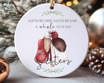 May include: A white ceramic Christmas ornament with a gold ribbon for hanging. The ornament features an illustration of two figures with the word "Sisters" and the text "A little bit crazy, a little bit loud, a whole lot of love."